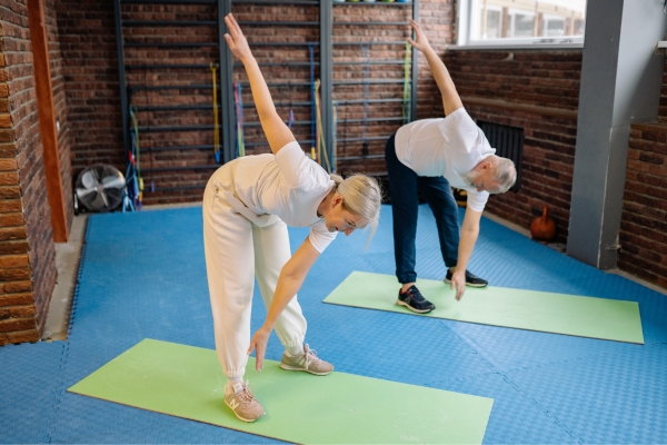 Ältere Frau und älterer Mann machen gemeinsam Dehnübungen auf Trainingsmatten in einem Fitnessraum.